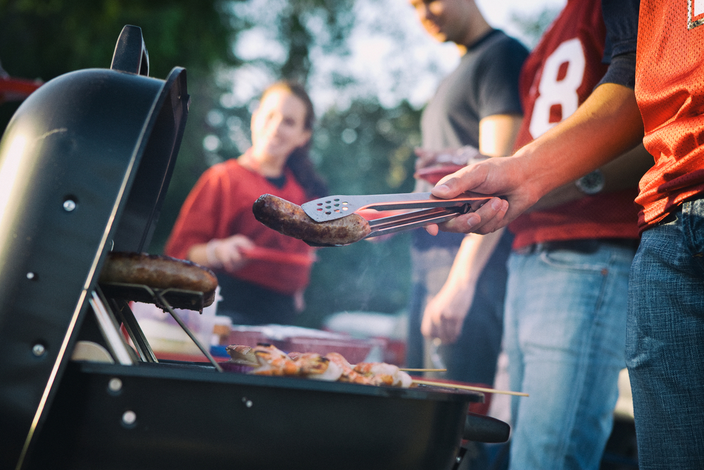A group of people surrounding a grill cooking brats at a tailgating party